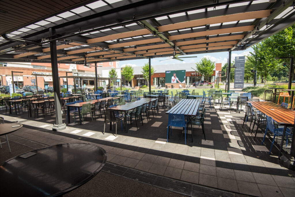 An outdoor dining area with numerous empty tables and chairs under a canopy. Surrounding buildings are visible in the background, along with green trees and a large screen displaying an image.