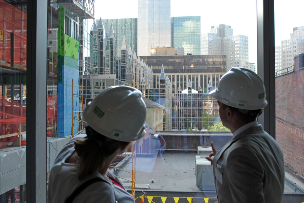 Two construction professionals in hard hats observing a building site through a large window. The view shows scaffolding and urban buildings under construction.