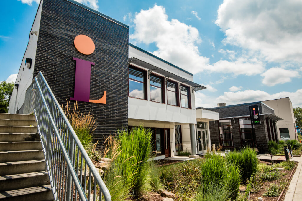 A modern building with large windows and the Carnegie Library of Pittsburgh logo on one side. The structure has stairs leading up to the entrance, surrounded by green grass and shrubs under a partly cloudy sky.