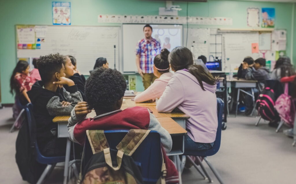 A classroom with students seated at desks facing a teacher in front of a projection screen. Students are listening attentively, with backpacks on some chairs.