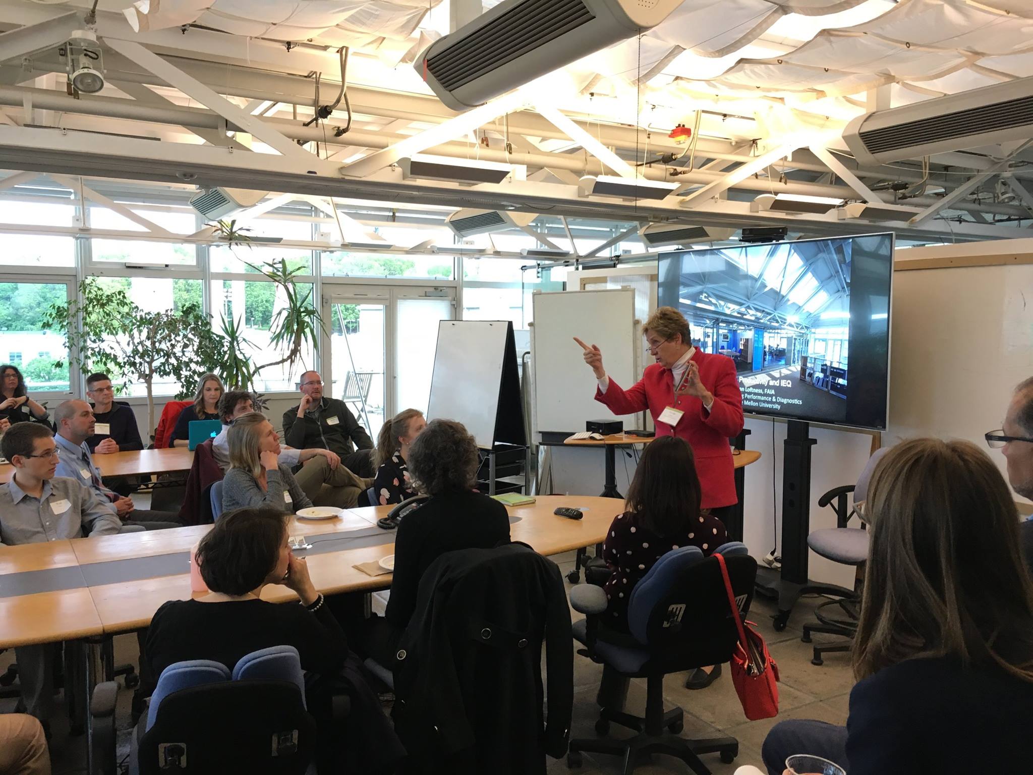 A group of people seated around tables in a conference room. A woman in a red blazer is speaking in front of a large screen displaying a presentation. Plants are visible through the windows.