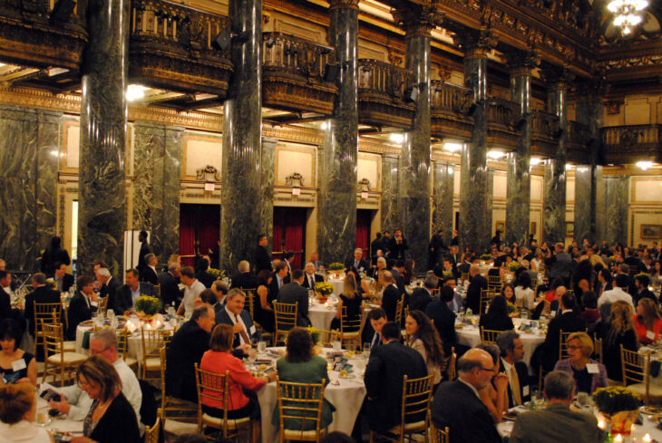 A large banquet hall filled with guests seated at round tables. Elegant decor features tall marble columns and decorative lighting. Attendees are engaged in conversation over dinner.
