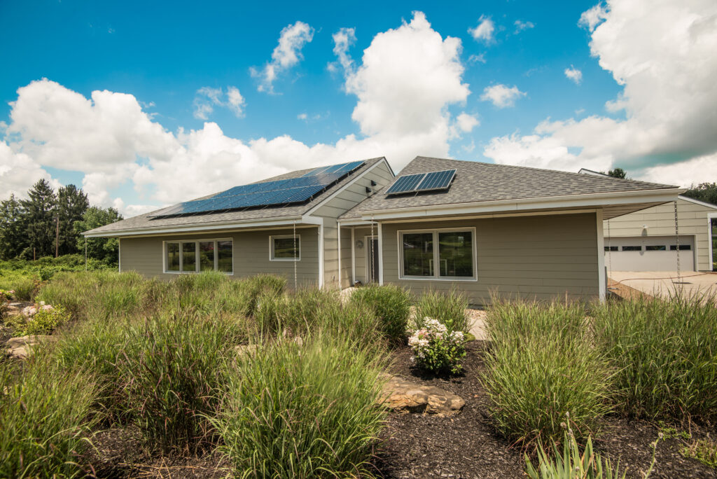 A modern building with solar panels on the roof, surrounded by grass and shrubs, under a partly cloudy sky. The building features a light-colored exterior and large windows.