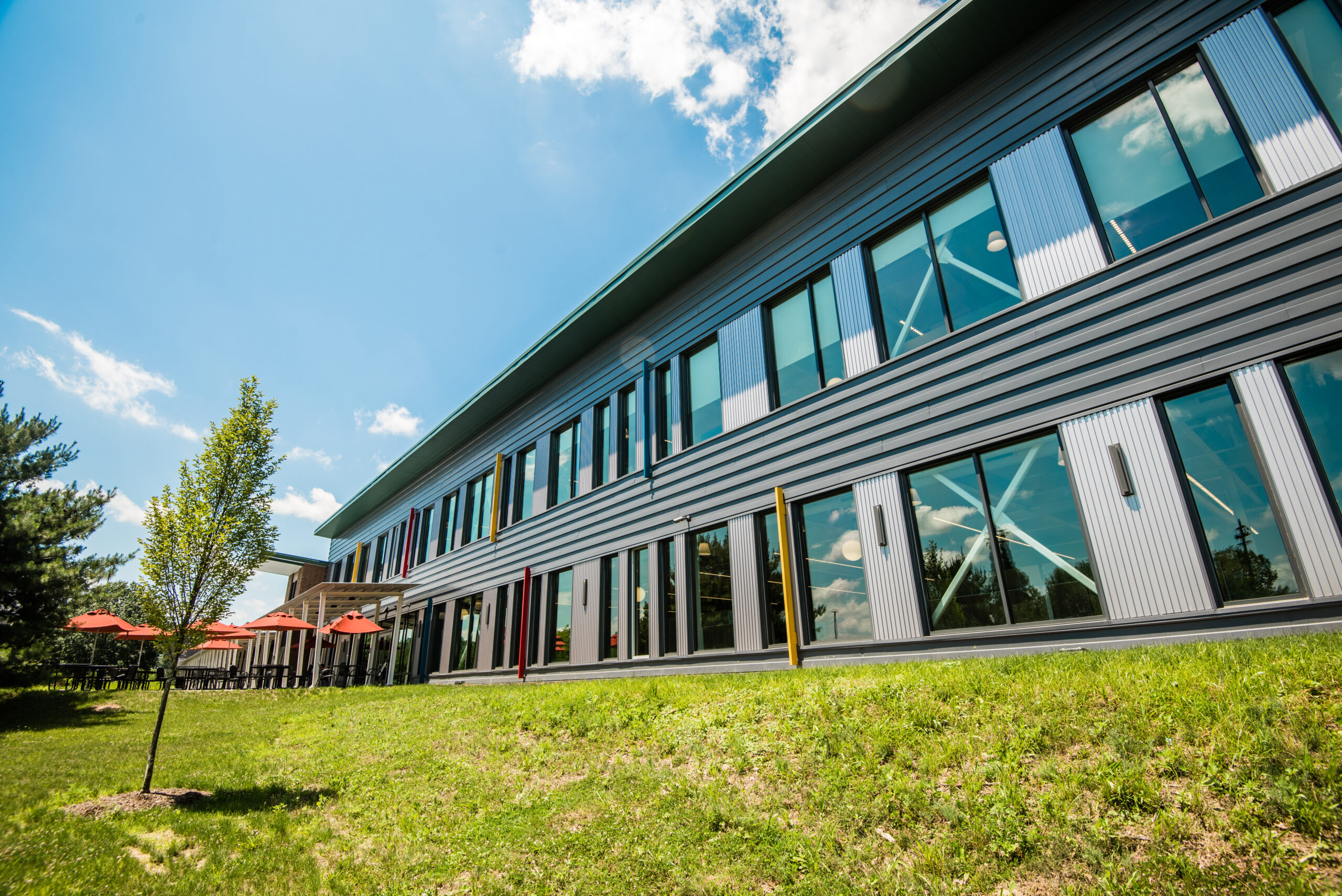 A modern building with large windows and a slanted roof, situated on a grassy area. Small trees and red umbrellas are visible in the foreground under a blue sky with clouds.