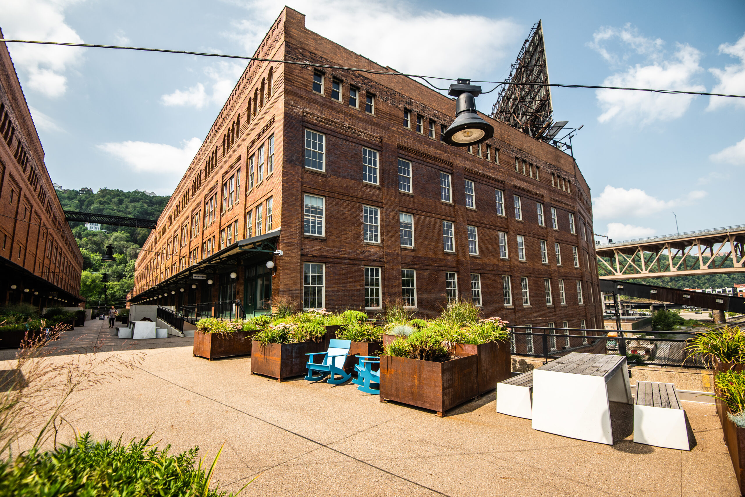 A red brick building with large windows surrounded by plants and outdoor seating. A metal bridge is visible in the background under a clear sky.