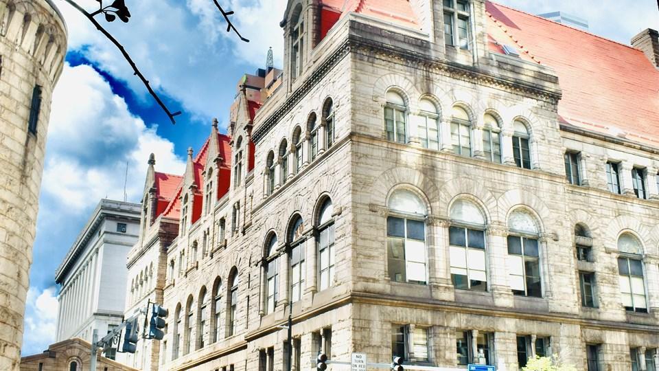 The exterior of the Allegheny County Courthouse, a historic stone building with large arched windows and a red roof, set against a cloudy sky. Street signs and lampposts are visible in the foreground.