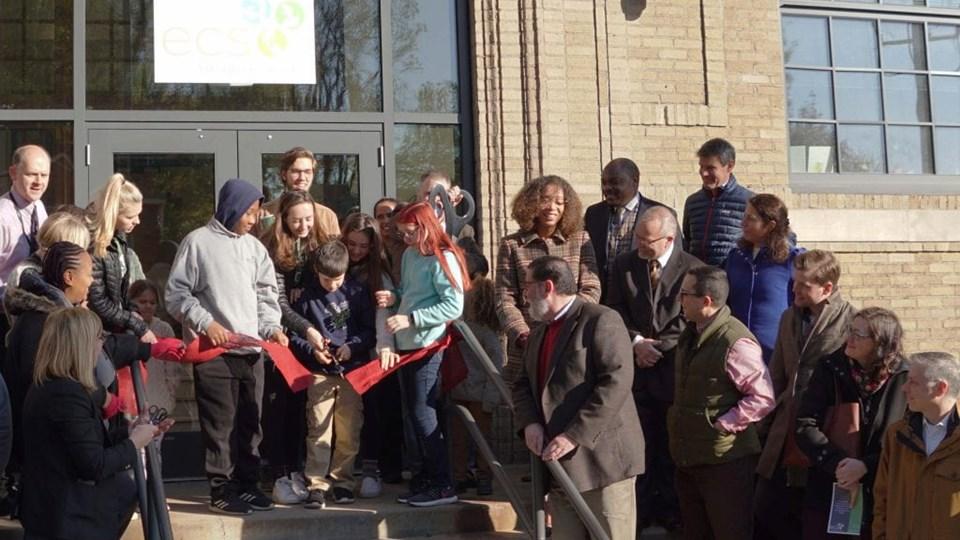 A group of people gathered at the entrance of the ECS new middle school building, participating in a ribbon-cutting ceremony. They are holding red ribbons and scissors.