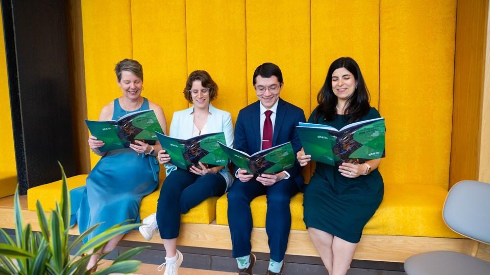 Four individuals seated on a yellow couch, each holding a magazine. They are looking at the magazines, wearing business attire.