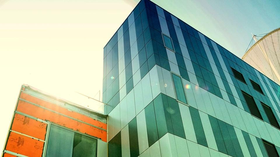 A modern building with a reflective facade composed of blue and gray panels, partially sunlight illuminated. The lower part shows an orange section under construction with exposed materials.