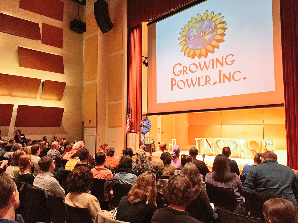 A speaker stands on stage in front of an audience, with a large screen displaying the logo of Growing Power, Inc. The room is filled with people seated and facing the stage.