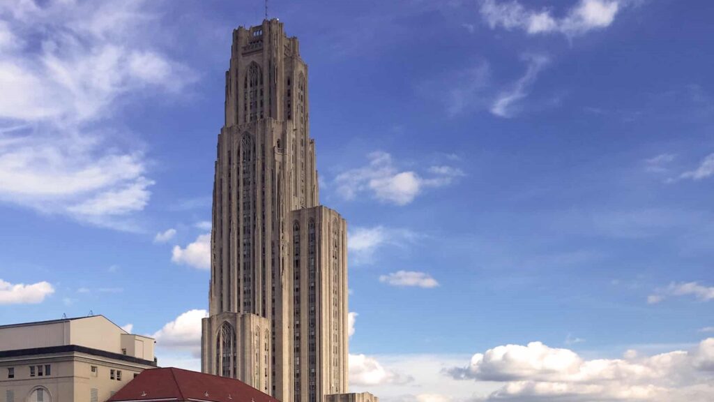 The Cathedral of Learning, a tall, iconic building with a distinctive architectural design, set against a blue sky with white clouds. The building is part of a city skyline, with a red roof visible in the foreground.