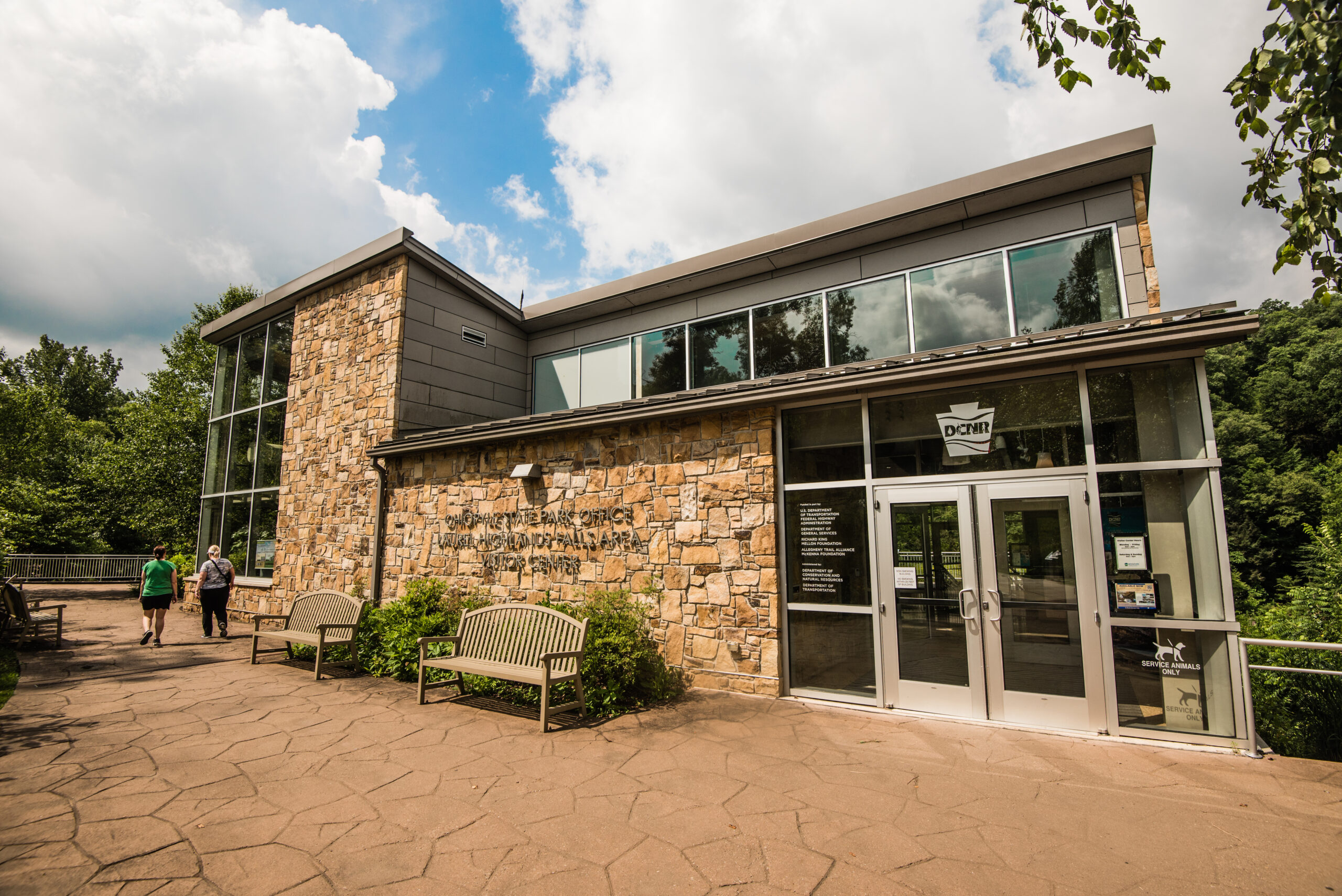 A modern building made of stone and glass with large windows. Two people walk towards the entrance, which has double doors and a sign. Surrounding the building are benches and trees, under a partly cloudy sky.