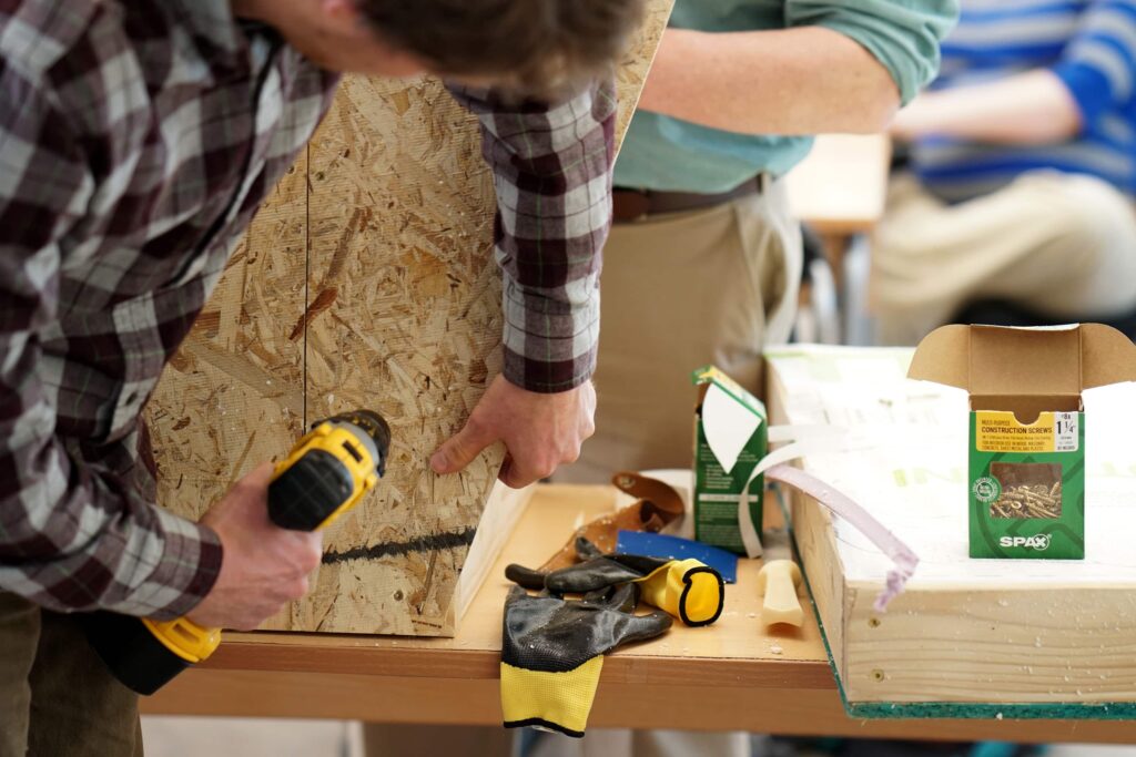A person using a power drill to fasten a piece of oriented strand board (OSB) on a workbench. Another person is handling materials nearby. Various tools and items, including gloves and a box of screws, are visible on the table.