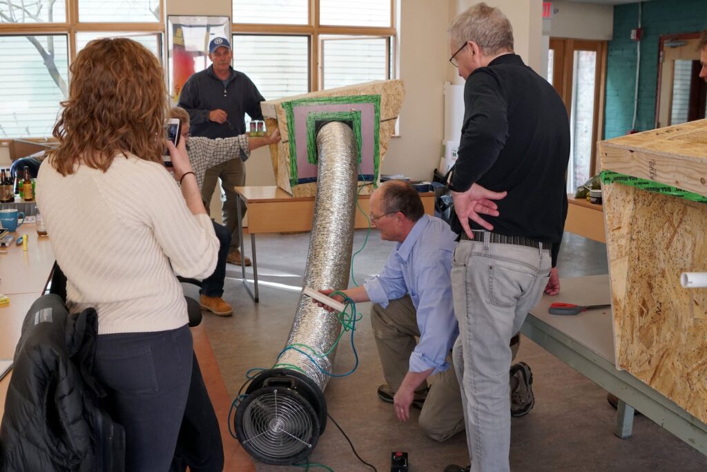 Passive House training. A group of people in a workshop setting working on a large duct. One person kneels beside the duct connecting equipment. Two others observe, and one takes a photo. Light comes through large windows in the background.