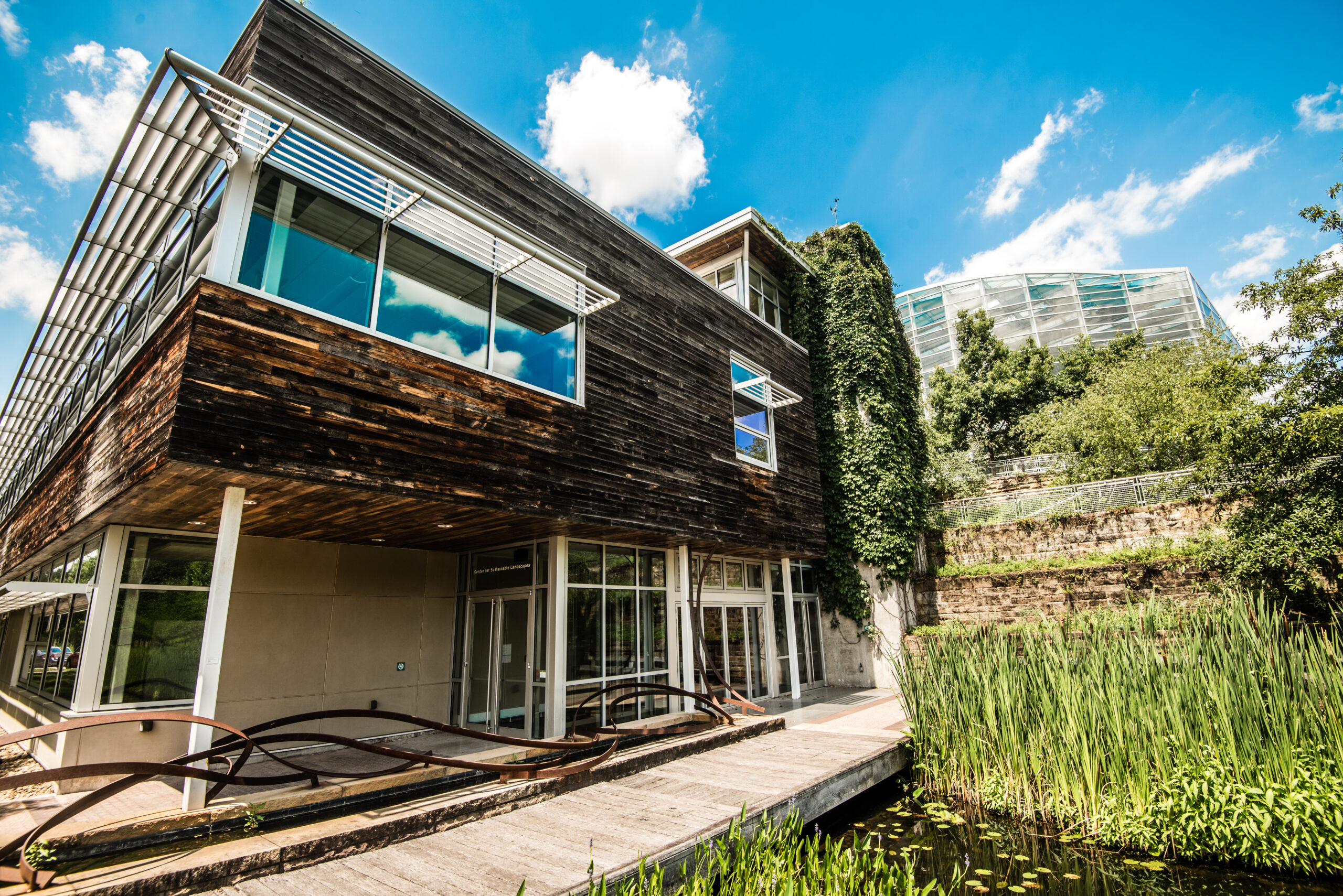 Center for Sustainable Landscapes exterior. A modern building featuring a wooden exterior with large glass windows, surrounded by greenery and a pond. A wooden walkway leads to the entrance, and a second structure is visible in the background.