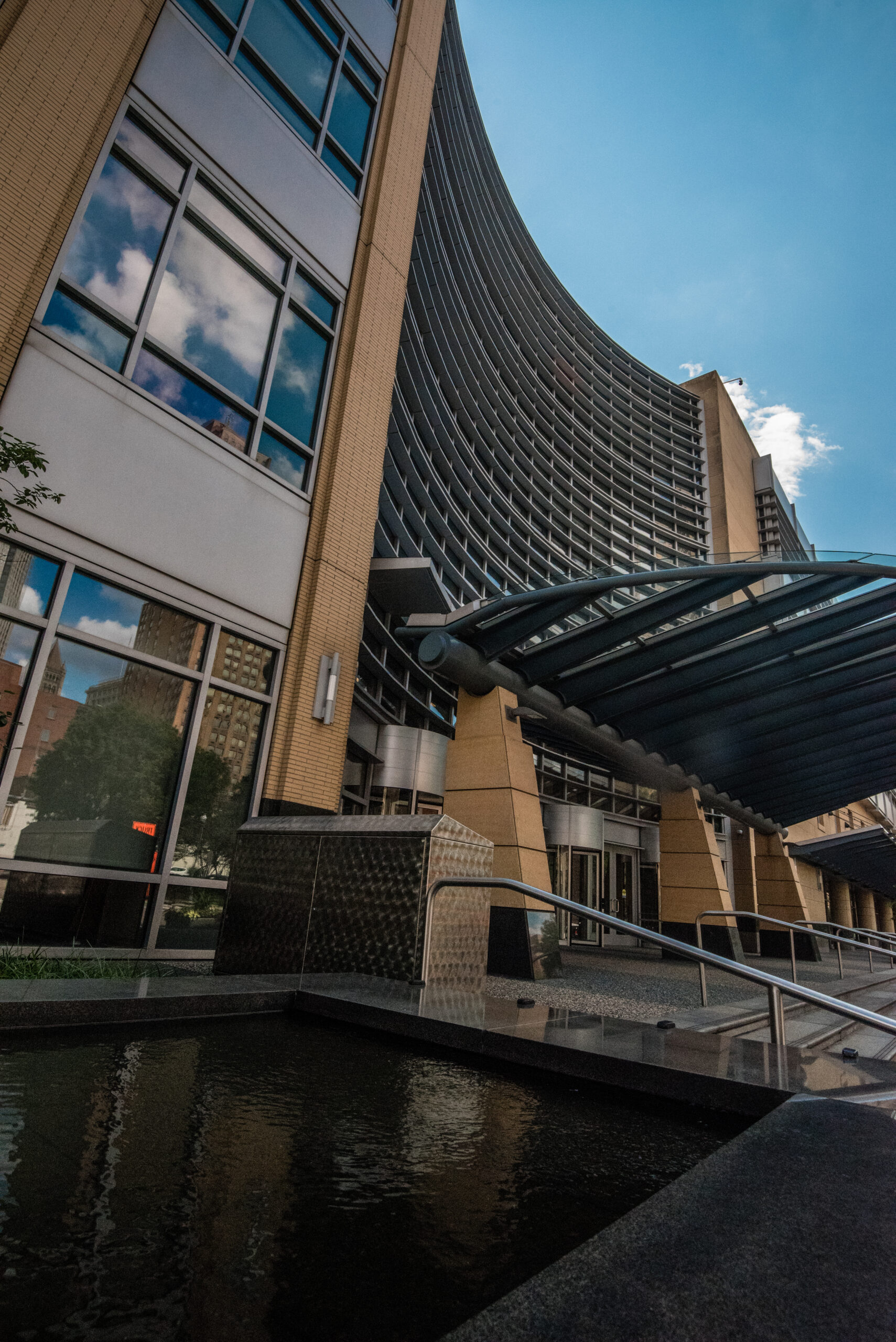 A modern building with a curved facade and large glass windows. The entrance is covered by a metal canopy, and a water feature is visible in the foreground. The sky is partly cloudy.
