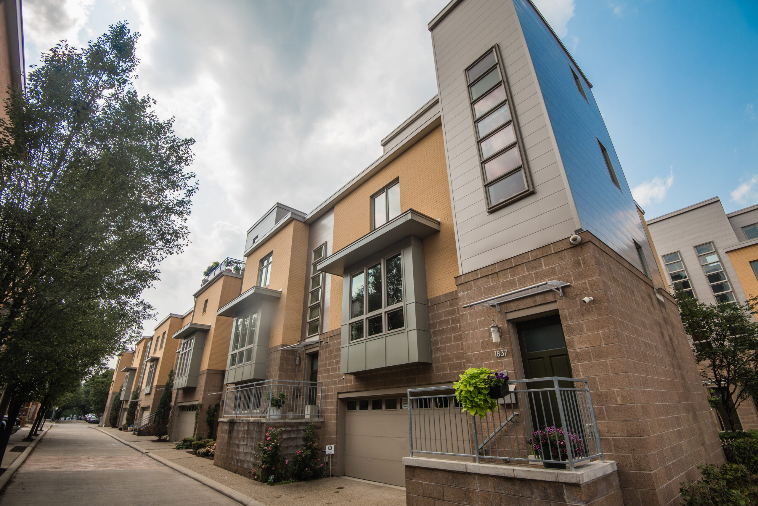 Modern townhouses lining a street with green trees on the left. The buildings feature a mix of materials, including stone and siding, with large windows and balconies.