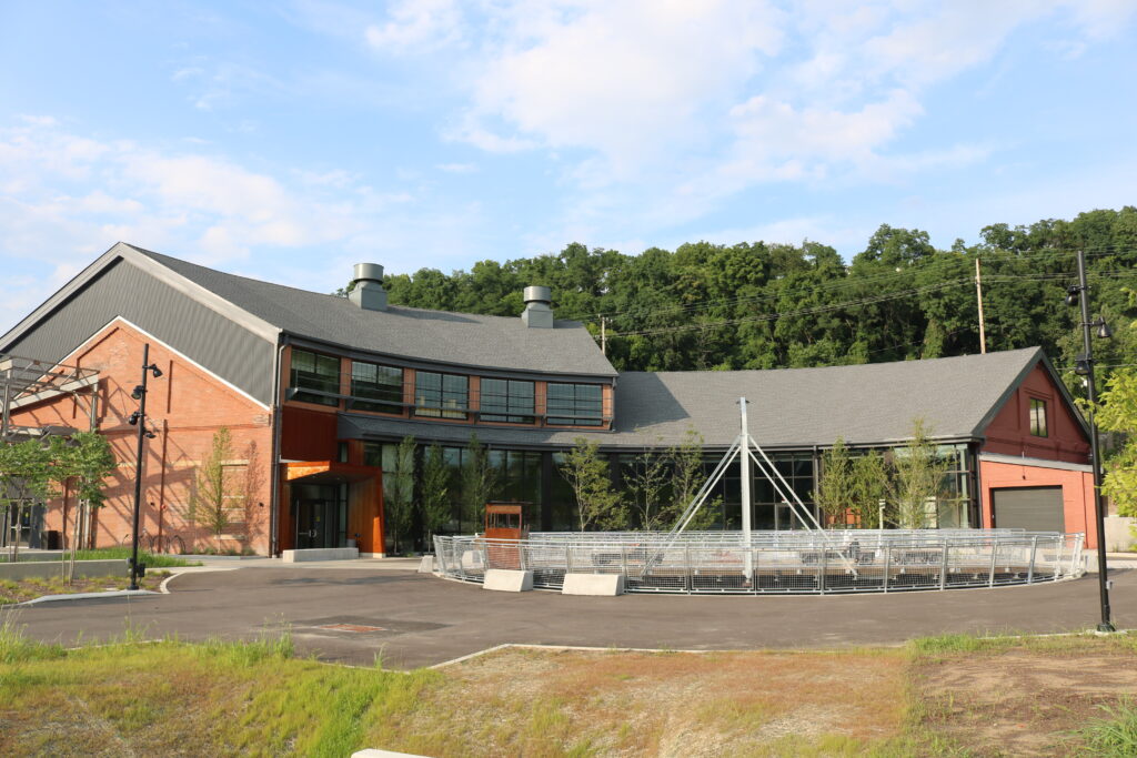 A modern two-story building with large windows and a gray roof, surrounded by greenery. In front, there is a circular paved area with benches and a large canopy structure. The setting includes trees and a blue sky in the background.