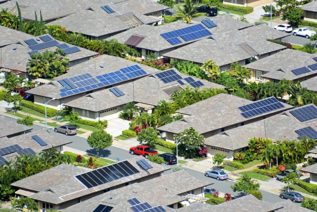 An aerial view of a residential neighborhood with multiple houses featuring solar panels on their roofs. Lush greenery and parked cars are also visible.
