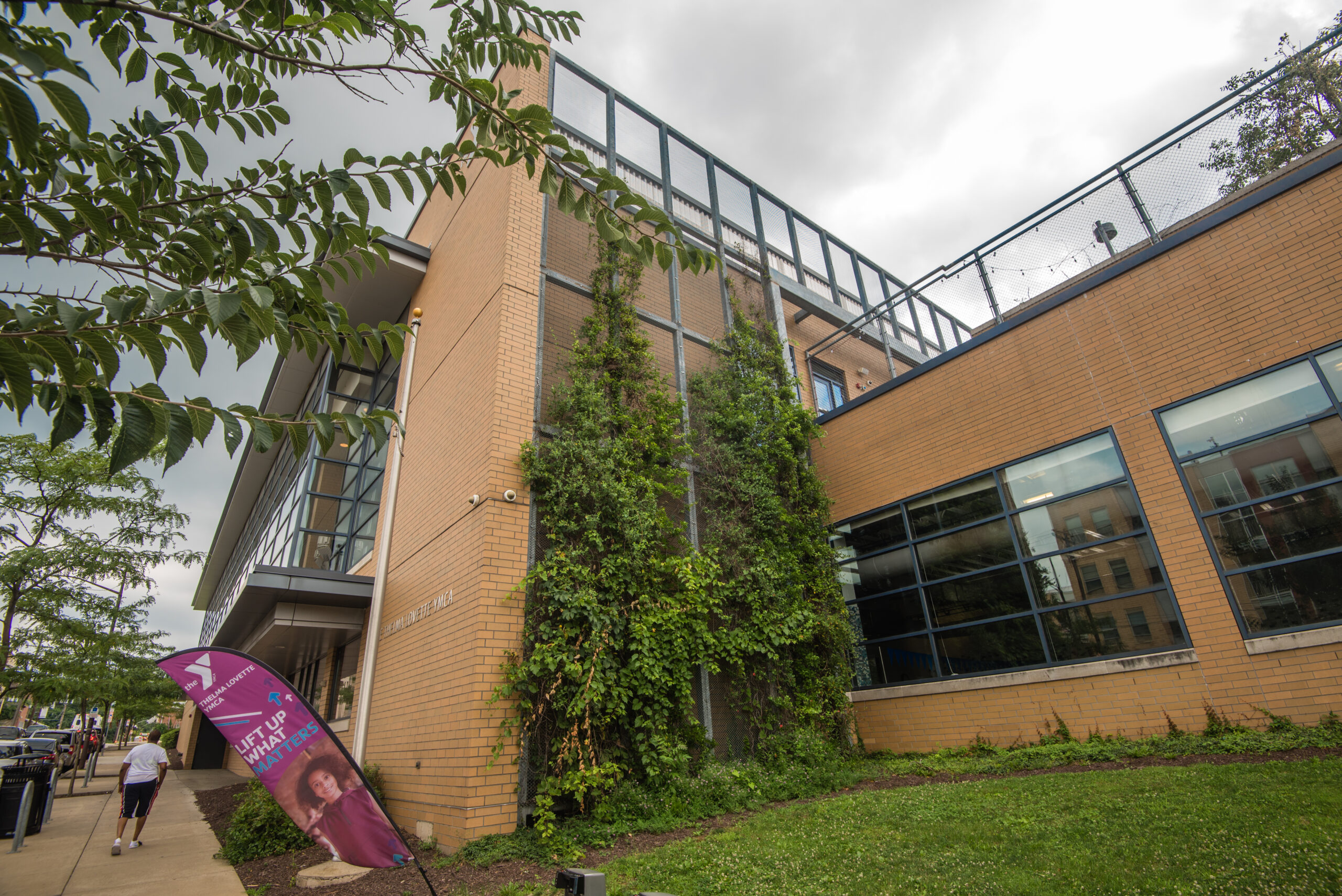 Exterior view of a modern building with large glass windows and greenery. A purple banner is displayed in front of the building. The scene is partly cloudy with trees nearby.