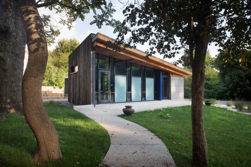 A modern building with a sloped roof, large windows, and wooden accents, surrounded by grass and trees. A concrete walkway leads to the entrance of the building.