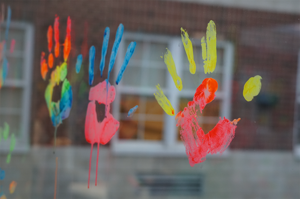 Colorful handprints in blue, pink, yellow, and orange painted on a glass window. The background shows a brick wall and a window.