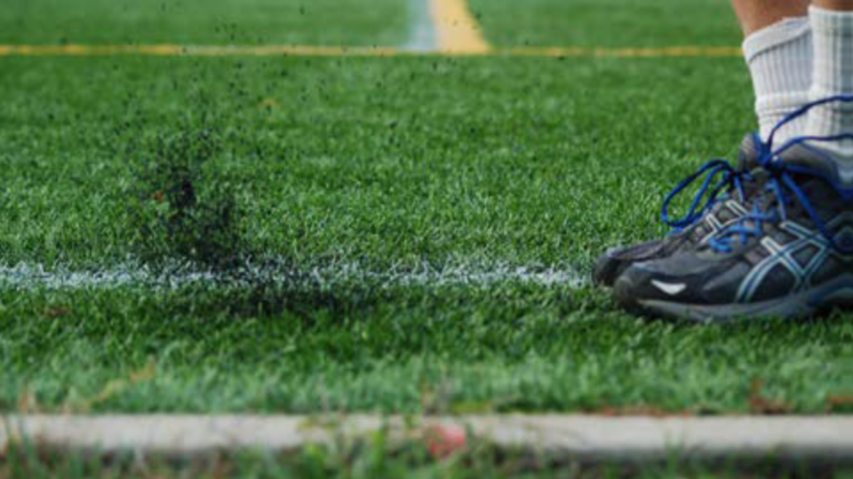 A close-up of a person's black athletic shoe on artificial turf, with small particles of rubber scattering from the foot, and a white line marking on the field visible in the background.
