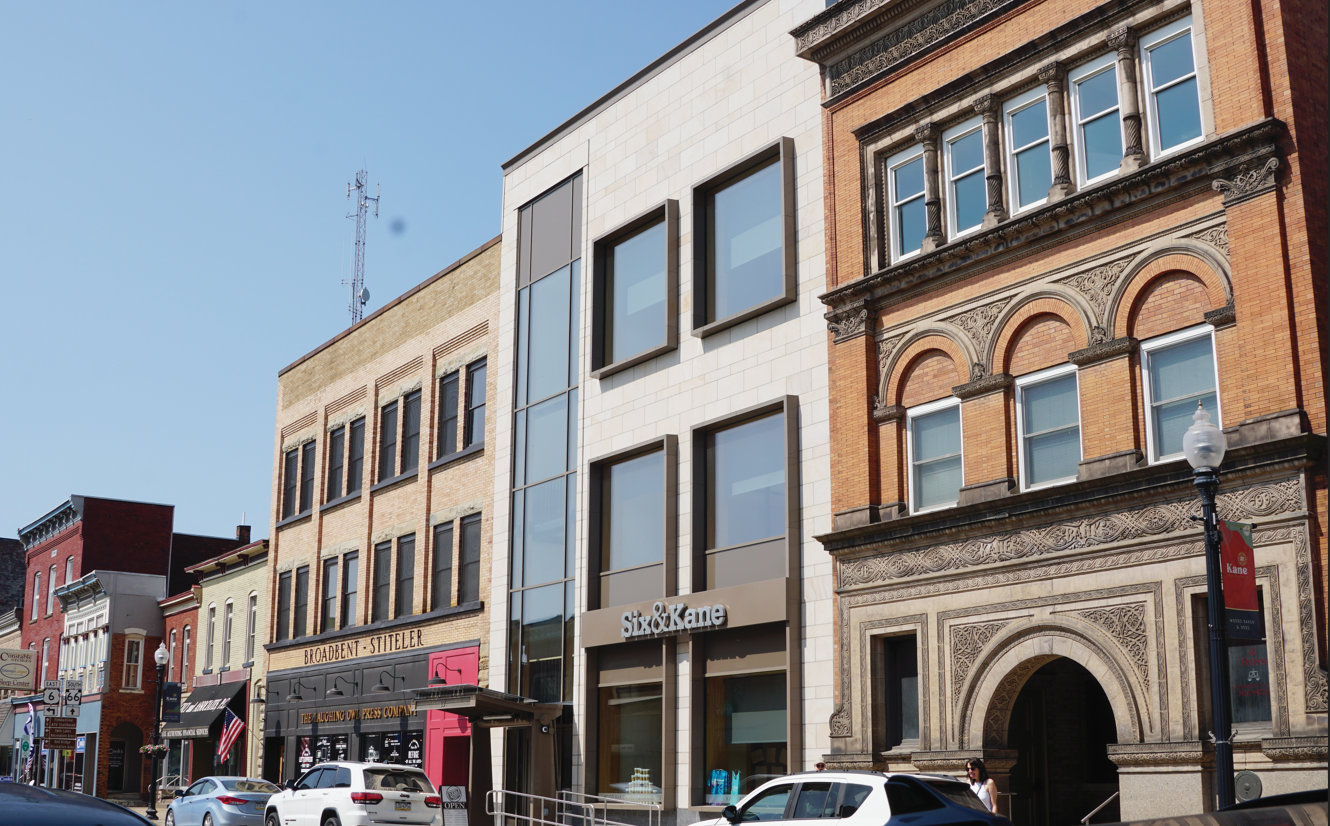 A streetscape showing a mix of architectural styles, including the Six&Kane Exterior, a modern building with large windows, and historic brick buildings. Cars are parked along the street, and a flag is visible on one building.